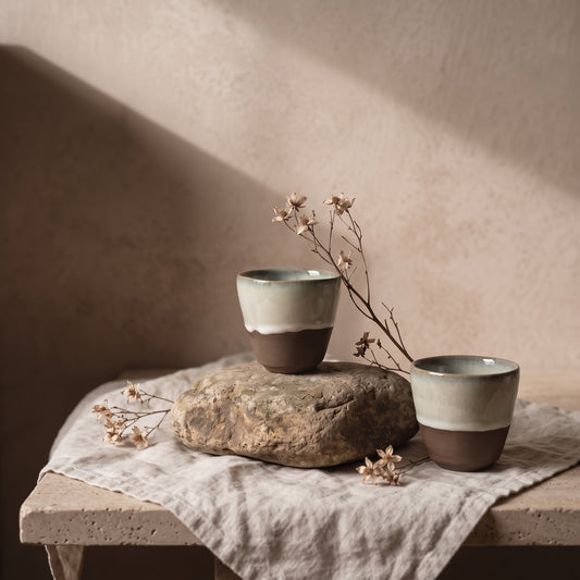 Two small ceramic cups, one with a green glaze on top and the other with a cream glaze on top, are resting on a large natural stone on a linen cloth, next to delicate dried flowers.