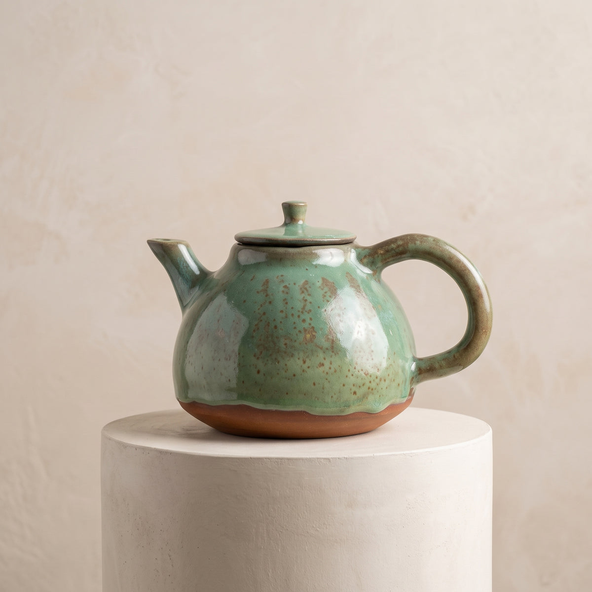 Green glazed ceramic teapot presented on round white display riser.