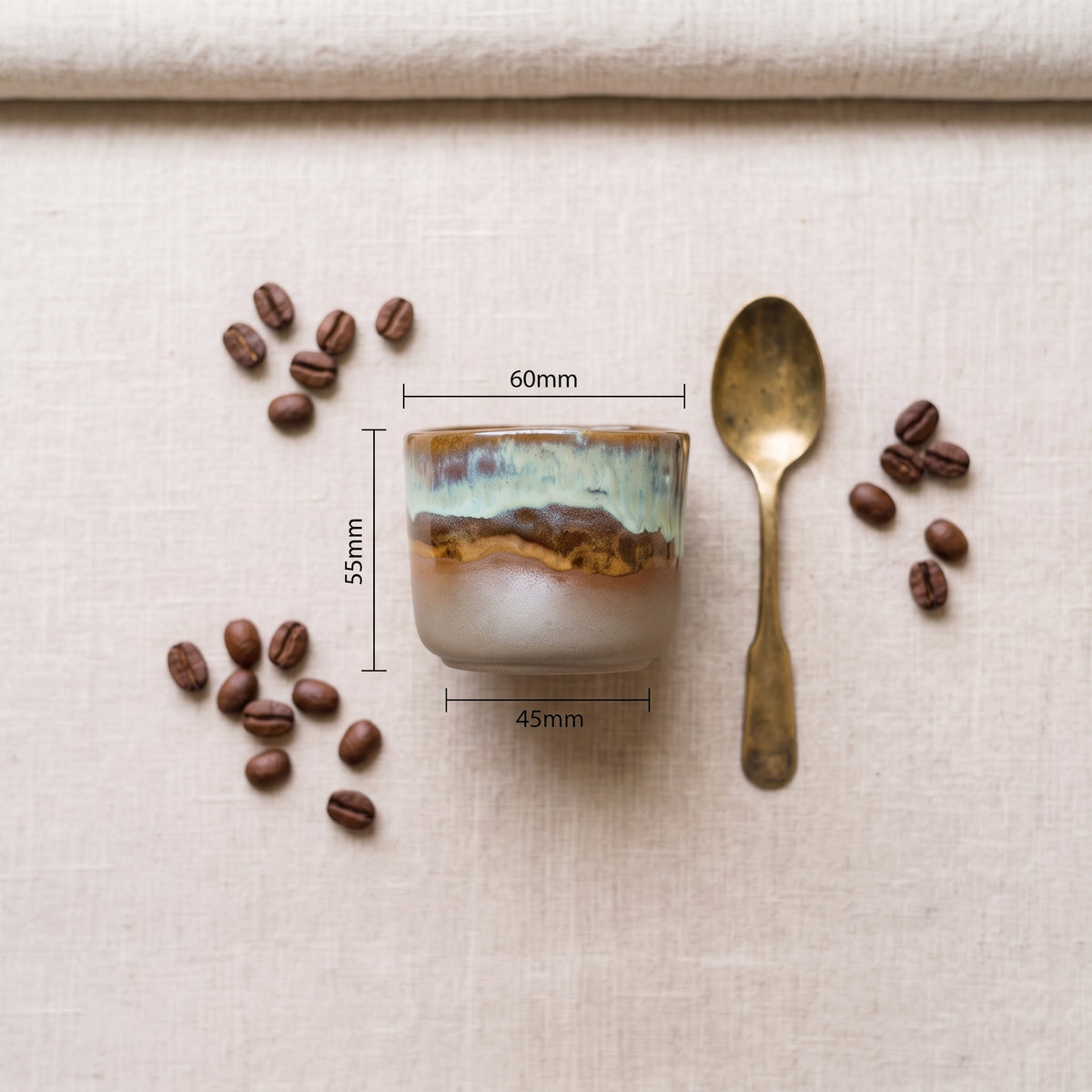 Overhead view dimension guide for a handmade ceramic espresso/shot cup with a brass spoon and scattered coffee beans on a linen tablecloth.
