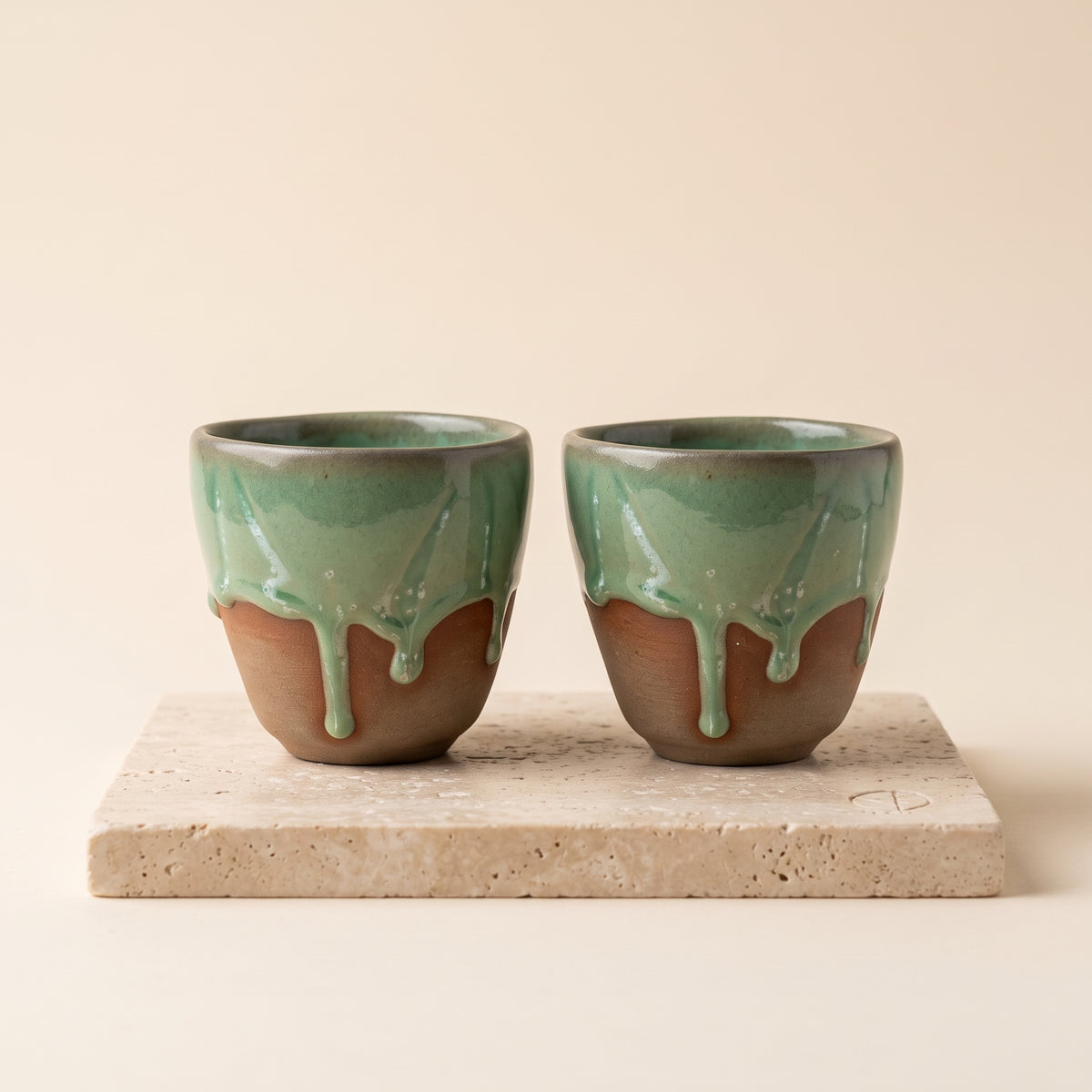 Pair of LyLceramics Olive Sip handmade ceramic espresso cups with dripping olive green glaze on a travertine plinth.