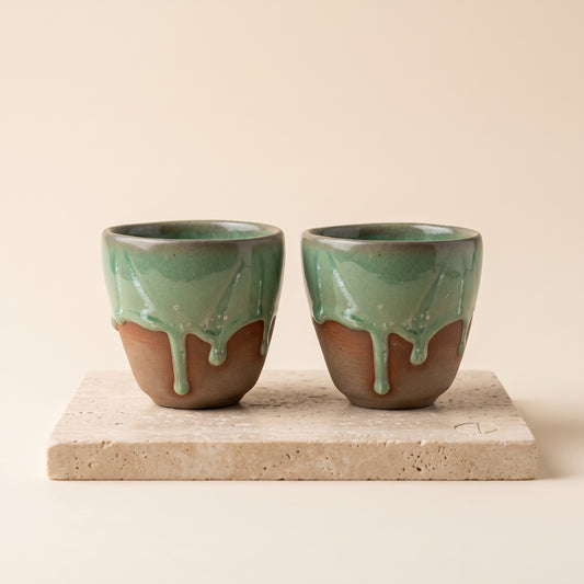 Pair of LyLceramics Olive Sip handmade ceramic espresso cups with dripping olive green glaze on a travertine plinth.