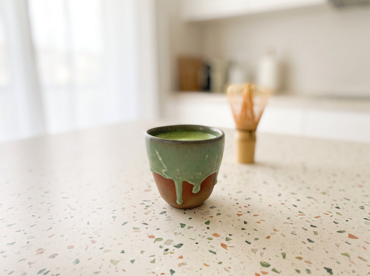 LyLceramics Olive Sip ceramic cup with bamboo whisk on a speckled stone countertop in a modern kitchen.