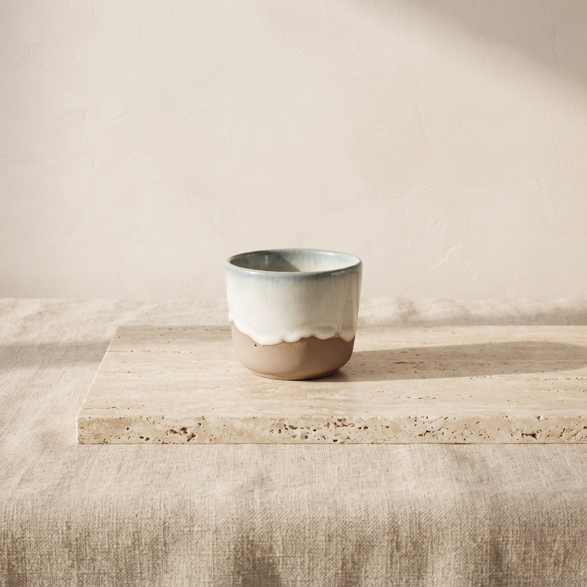 Handmade stoneware tumbler centered on a wrinkled textured linen tablecloth in soft daylight.