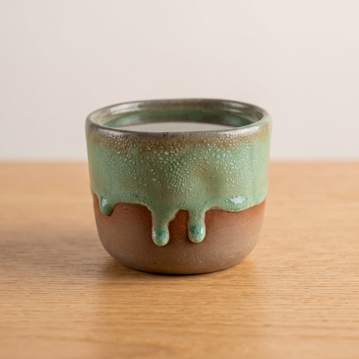 Front view of a handmade olive green stoneware shot glass resting on a wooden tabletop.