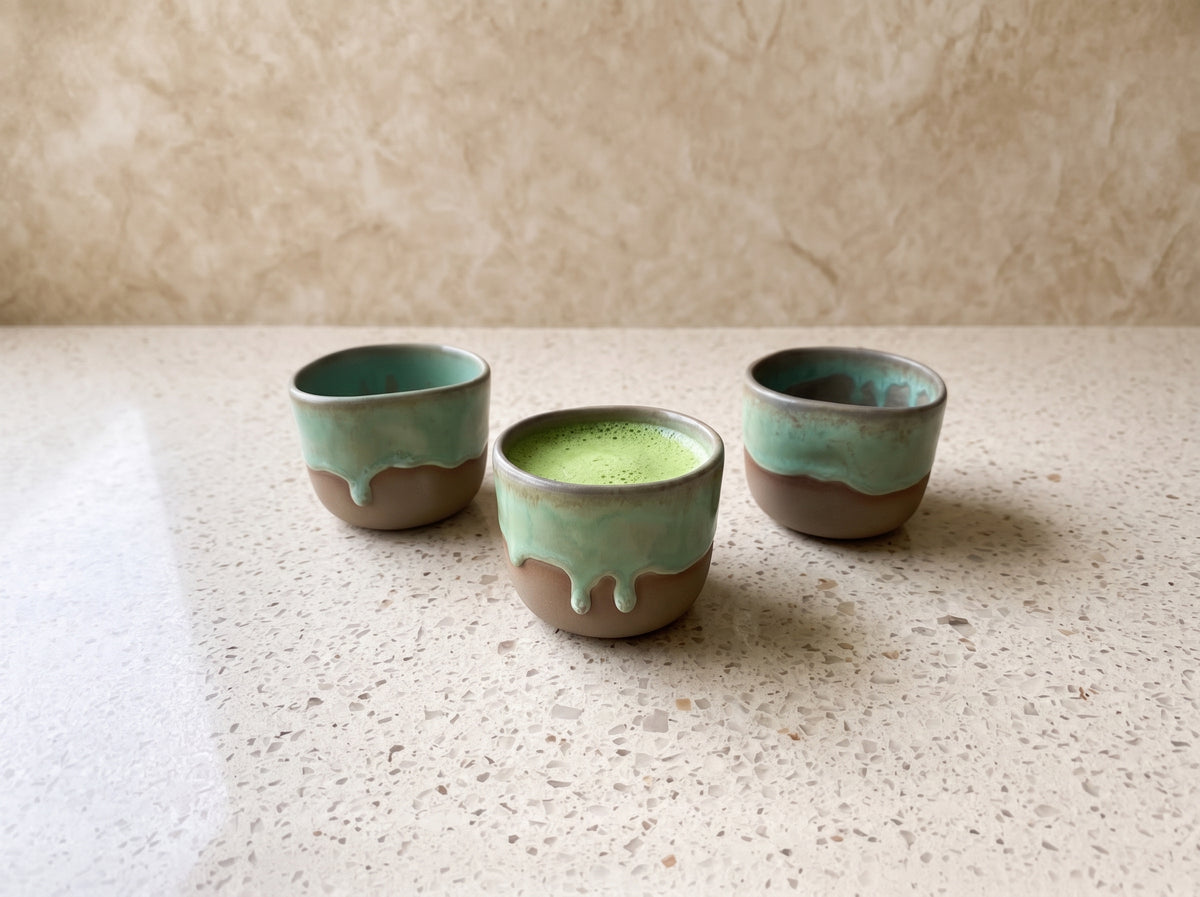 Three handmade olive green ceramic shot glasses arranged on a speckled stone countertop.