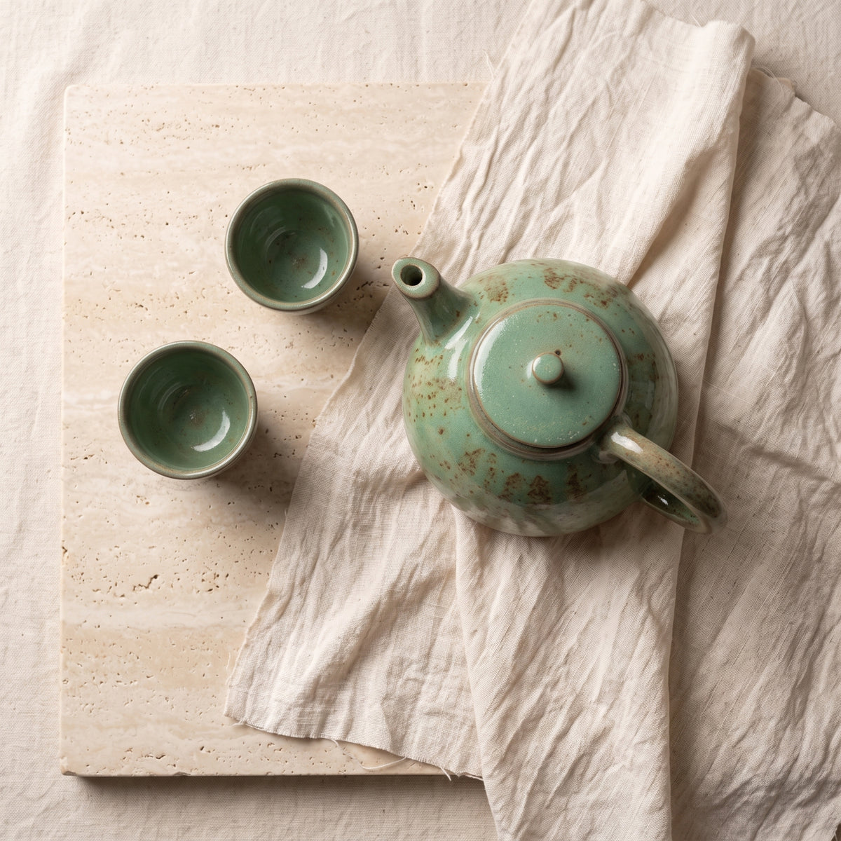 Overhead flat lay of ceramic teapot and cups styled on linen cloth and wood board.