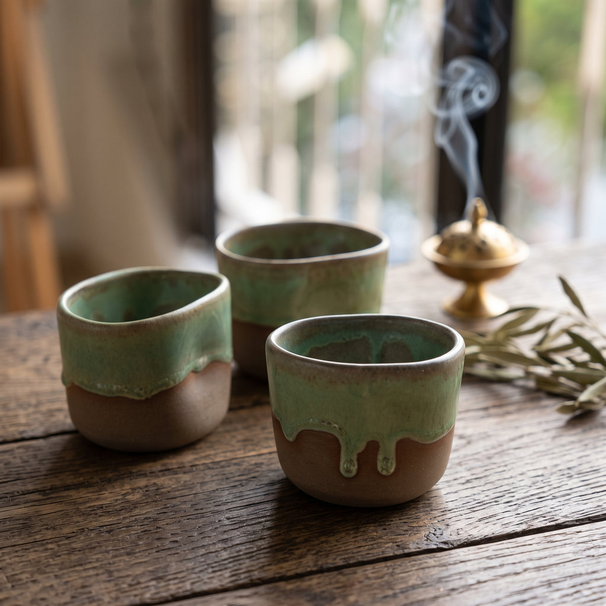 Three small, rustic ceramic cups with green and brown glazes are arranged on a dark wooden table, beside a burning brass incense burner and a sprig of dried leaves.